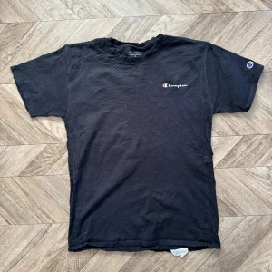Svart t-shirt från Champion - Svart t-shirt från Champion med klassisk logga på bröstet och ärmen. Enkel och stilren design, perfekt till jeans eller shorts. Tillverkad i mjuk bomull och har en normal passform med korta ärmar.