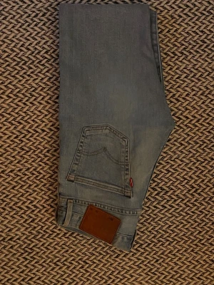 Levis 501 Premium -W26 -L30 - Säljer ett par ljusblå jeans med raka ben och klassisk femficksdesign. Jeansen har en brun patch bak i midjan och en liten röd detalj vid bakfickan. Perfekta för en avslappnad och trendig look.