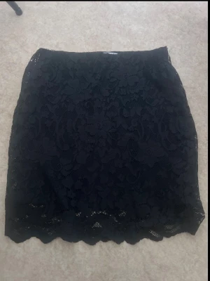 Svart spetskjol med blommönster - Snygg svart kortkjol i spets med blommigt mönster och vågig nederkant. Kjolen har en feminin och elegant look, perfekt att styla med en enkel topp eller stickad tröja. Materialet är mjukt och följsamt för en bekväm känsla.