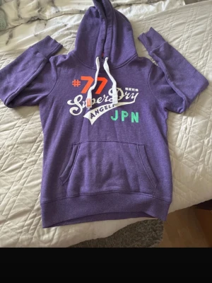 Lila Superdry hoodie med tryck - Säljer en lila hoodie från Superdry med stor vit logga, orange #77, grönt JPN-tryck och vita snören i huvan. Hoodien har känguruficka och ribbade muddar. Perfekt för en avslappnad och sportig stil.