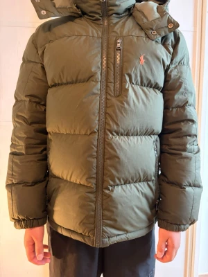 Olivgrön Ralph lauren med huva - Säljer en olivgrön dunjacka från Polo Ralph Lauren med huva och dragkedja framtill. Jackan har en bröstficka med dragkedja, två sidofickor och den klassiska loggan broderad på bröstet. Perfekt för kalla vinterdagar och riktigt skön att bära. Priset är inte skrivet i sten!