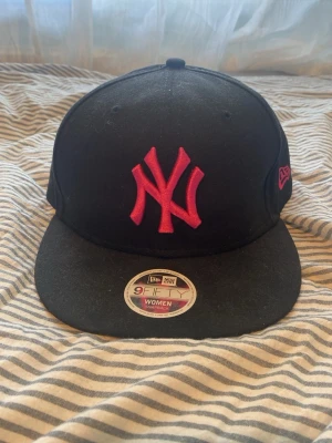 Svart New Era keps med rosa NY-logga - Snygg svart snapback-keps från New Era med rosa broderad NY-logga framtill och rosa New Era-logga på sidan. Klassisk platt skärm och justerbar baksida. Passar perfekt för dig som vill ha en sportig och trendig stil.