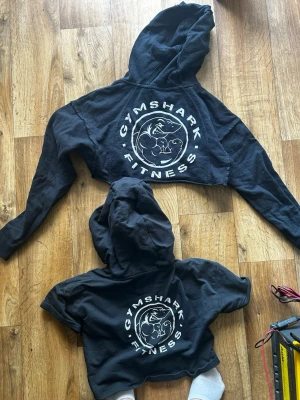 Svart cropped hoodie från Gymshark - Säljer en svart cropped hoodie från Gymshark med stort vitt tryck på bröstet där det står 'GYMSHARK FITNESS' och en muskulös haj i mitten. Hoodien har huva och långa ärmar, perfekt för gymmet eller chill. Materialet känns mjukt och bekvämt. Den andra är kortärmad säljer båda tillsammans för 200kr
