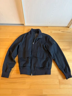 J.Lindeberg Men's Bomber Jacket - This is a men's bomber jacket from J.Lindeberg. The jacket is black, features a zip-up front, two large flap pockets on the front, ribbed cuffs and hem, and a stand collar. The left sleeve has a rubber J.Lindeberg logo patch.