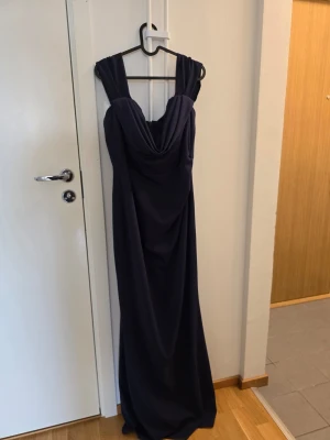 Mörkblå långklänning med drapering - Snygg mörkblå långklänning med breda axelband och draperad urringning. Klänningen har en elegant, figurnära passform och faller mjukt ner till golvet. Perfekt för dig som vill ha en stilren och klassisk look.