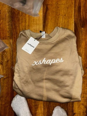 Beige tröja från xshapes - Snygg beige tröja från xshapes med vit broderad logga på bröstet. Klassisk passform med ribbade muddar vid ärmslut och nederkant. Perfekt för en chill och stilren look.