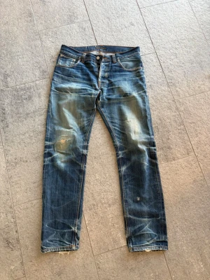 Blå slitna jeans med raka ben - Säljer ett par blå jeans med slitningar och tvättade detaljer. Jeansen har raka ben, klassisk femficksmodell och synliga slitningar på knän och bakfickor. Materialet är denim i bomull och passformen är avslappnad. Perfekta för en avslappnad streetstil.