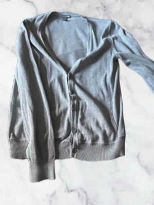 Grå cardigan med knappar - Stilren grå kofta med v-ringning och knäppning framtill. Tillverkad i ett tunt, mjukt material och har ribbade muddar vid ärmslut och nederkant. Perfekt att slänga över en t-shirt för en enkel look.