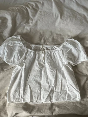 Vit topp med puffärm - Söt vit topp med puffiga korta ärmar och liten knytning framtill. Toppen har en luftig och lätt känsla, perfekt till sommaren. Passar till både jeans och kjol för en trendig look.