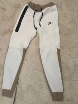 Vita och beige mjukisbyxor från Nike - Säljer ett par snygga vita mjukisbyxor från Nike med beige detaljer och svarta snören i midjan. Byxorna har en svart dragkedja längs ena benet och muddar vid bensluten. Perfekta för chill eller träning!