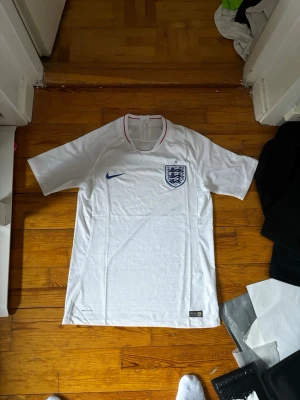 Vit England fotbollströja - Säljer en vit England fotbollströja med blå Nike-logga och landslagets emblem på bröstet. Tröjan har korta ärmar, rund hals med röd detalj och är tillverkad i ett lätt och ventilerande material. Perfekt för fotbollsträning eller match.