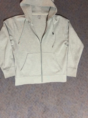 Ljusgrå hoodie från Polo Ralph Lauren - Ljusgrå hoodie med dragkedja från Polo Ralph Lauren. Klassisk design med huva, dragsnören och två fickor framtill. Broderad logga i mörkblått på bröstet. Mjuk bomullsblandning som känns skön mot huden.