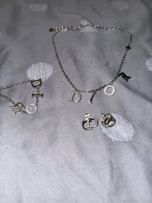 Silverfärgat halsband med DIOR-bokstäver - Snyggt halsband i silverfärg med hängande bokstäver som bildar ordet DIOR. Bokstäverna är dekorerade med små glittrande stenar för extra bling. Perfekt accessoar för att lyfta din outfit och visa din stil.