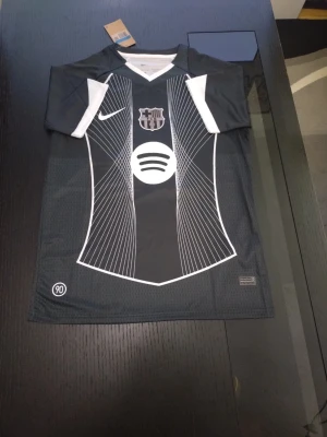 Svart Barcelona matchtröja Nike - Snygg svart Barcelona matchtröja från Nike med vita detaljer och grafiskt mönster på framsidan. Tröjan har klubbmärke, Nike-logga och Spotify som sponsortryck. Korta ärmar och V-ringad hals med vit kant. Perfekt för dig som älskar fotboll och vill sticka ut på planen.