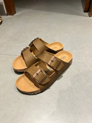 Bruna Birkenstock sandaler med spännen - Säljer ett par klassiska bruna Birkenstock sandaler i storlek 41. De har två breda remmar i mocka med justerbara metallspännen och en bekväm korksula. Perfekta för dig som gillar enkel och tidlös stil. Dem är du inte äkta därför billigare, kan skicka i augusti först/6