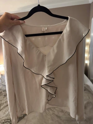 Ljusbeige blus med volangdetalj - Supersöt ljusbeige blus med volang längs ringningen och framtill, samt svarta kontrastsömmar. Blusen har v-ringning och långa ärmar. Materialet känns lätt och silkeslent, perfekt för en trendig och chic look.