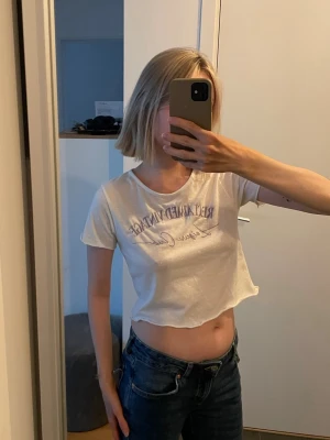 Vit croppad t-shirt  - Säljer en vit croppad t-shirt från Reclaimed Vintage i storlek M med broderat tryck framtill i lila text. T-shirten har korta ärmar och en avslappnad passform. 10 kr!