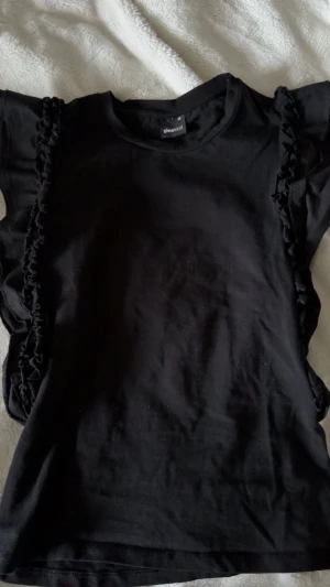Svart t-shirt med volangdetaljer - Svart t-shirt från Gina Tricot med korta ärmar och volangdetaljer längs sidorna. T-shirten har en klassisk rund halsringning och är tillverkad i mjuk bomull. Perfekt för en trendig och stilren look.