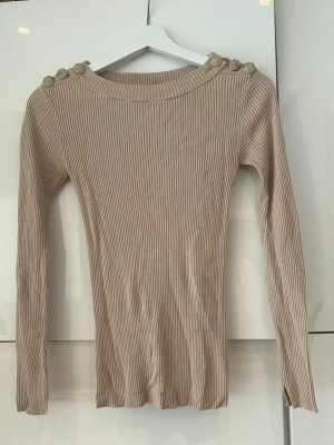 Beige ribbad topp från Hunkydory - Säljer en beige ribbad långärmad topp från Hunkydory. Toppen har dekorativa knappar på axlarna och en rund halsringning. Materialet är mjukt och stretchigt, vilket gör den både bekväm och snygg.