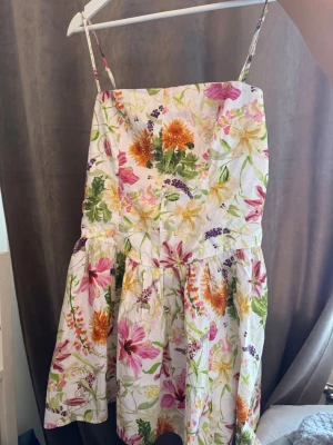 Blommig sommarklänning med smala band från hm - Söt kortklänning de med smala axelband och färgglatt blommönster i rosa, orange, lila och grönt på vit botten. Klänningen har en luftig passform och volang nedtill, perfekt för varma dagar. Tillverkad i bomull för en skön och sval känsla.