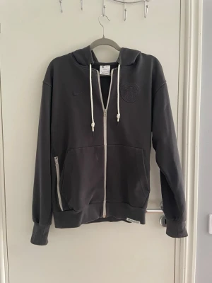 Mörkgrå zip hoodie med NBA-detalj - Snygg mörkgrå hoodie med dragkedja och vita snören. Har huva, fickor framtill och en NBA-logga på ärmen. Diskret Nike-logga och broderad emblem på bröstet. Perfekt för dig som gillar sportig stil och streetwear.