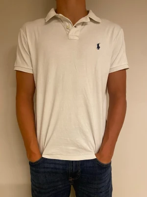 Vit pikétröja från Polo Ralph Lauren - Klassisk vit pikétröja från Polo Ralph Lauren med blå logga på bröstet. Tröjan har korta ärmar, krage och knappar vid halsen. Modellen är custom slim fit och tillverkad i mjuk bomull som känns skön mot huden.