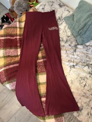 Vinröda Harvard yoga pants  - Säljer ett par vinröda leggings med hög midja och utsvängda ben. På ena höften står det 'Harvard University' i vitt tryck. Mjukt och stretchigt material som sitter skönt på kroppen. Perfekta för en chill och avslappnad stil.