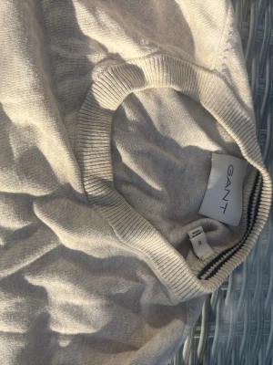 Beige stickad tröja från GANT - Klassisk beige stickad tröja från GANT med rund halsringning och diskret logga broderad på bröstet. Tröjan har långa ärmar och ribbade muddar vid hals, ärmslut och nederkant. Perfekt för lager på lager och enkel att matcha.
