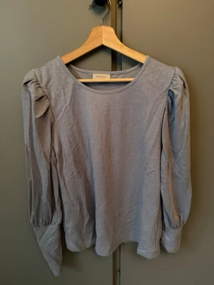 Ljusgrå långärmad topp med puffärm - Snygg ljusgrå topp med rund halsringning och långa puffärmar. Tillverkad i mjukt bomullsmaterial som känns skönt mot huden. Perfekt att styla med jeans eller kjol för en trendig look.