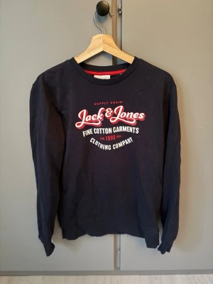 Marinblå sweatshirt från Jack & Jones - Cool marinblå sweatshirt från Jack & Jones med tryck i vitt och rött på bröstet. Tröjan har rund halsringning, långa ärmar och är gjord i mjuk bomull. Perfekt för en avslappnad streetwear-look.