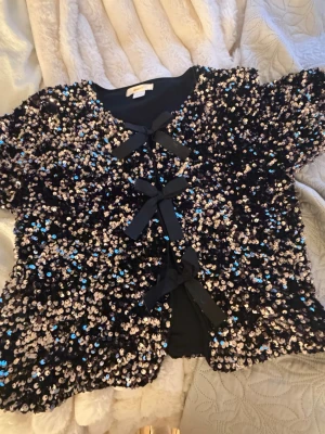 Paljettkofta från Gina Tricot - Glittrig svart kofta från Gina Tricot täckt med silver- och blå paljetter. Koftan har korta ärmar och knyts framtill med tre svarta rosetter. Perfekt för dig som vill sticka ut och älskar detaljer som glittrar.
