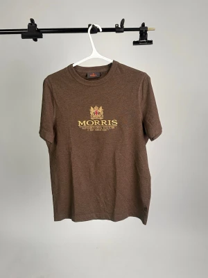 Brun t-shirt från Morris - Brun t-shirt från Morris med tryck i gult och rött på bröstet där det står 'Morris Sporting Club'. Klassisk rund halsringning och korta ärmar. Perfekt för dig som gillar stilrena och tidlösa plagg.