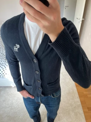 Mörkblå cardigan med knappar från Abercrombie&Fitch - Snygg mörkblå cardigan med knäppning framtill och två fickor. Tröjan har ribbade muddar och en vit broderad logga på bröstet. Perfekt att slänga över en t-shirt för en clean och avslappnad look.