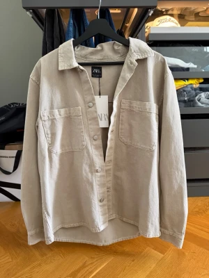 Beige overshirt från Zara - Stilren beige overshirt från Zara med två stora bröstfickor och klassisk krage. Skjortan har knappar framtill och är tillverkad i ett mjukt bomullsmaterial. Perfekt för lager på lager och en avslappnad look.