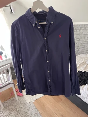 Mörkblå skjorta från Ralph Lauren - Klassisk mörkblå skjorta från Ralph Lauren med knappkrage och vit knappstängning framtill. Skjortan har lång ärm och den ikoniska röda logotypen broderad på bröstet. Perfekt till jeans eller chinos för en clean look.