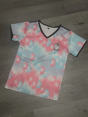 Belgien fotbollströja  - Säljer en kortärmad t-shirt med v-ringning och svarta detaljer vid hals och ärmslut. T-shirten har ett grafiskt mönster i rosa, blått och vitt med tunna ränder och halv-cirklar. Tillverkad i syntetmaterial och har en sportig vibe. 
