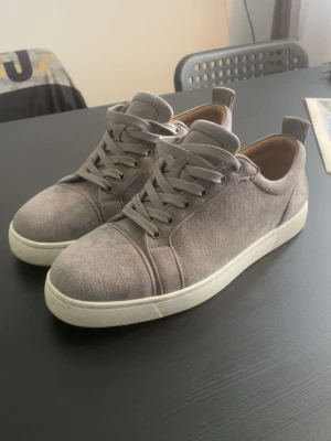 Gråa Christian Louboutin Sneakers - Snygga grå sneakers från christian louboutin i mocka med vita sulor och matchande grå skosnören. Klassisk låg modell med rund tå och diskreta sömmar för en clean look. Perfekta för dig som gillar stilrena och enkla skor. Bag & Org sula medföljer
