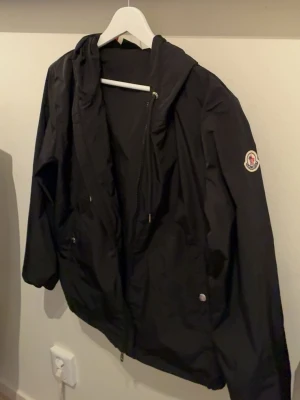 Svart vindjacka med huva Moncler - Snygg svart vindjacka från Moncler med huva och dragkedja framtill. Jackan har Moncler-logga på ärmen, två fickor med knappstängning och justerbar nederkant. Perfekt för blåsiga dagar och enkel att matcha till olika outfits. Jackan är i storlek 1, säljer på grund att jag vuxit ur den. För fler frågor eller funderingar tveka inte på att skriva!