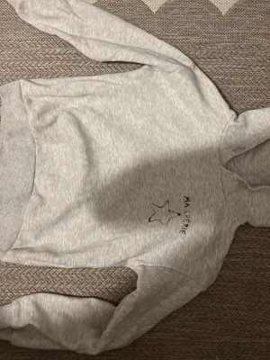 Ljusgrå hoodie med tryck Ma Chérie - Ljusgrå hoodie med svart texten 'MA CHÉRIE' och en stjärna tryckt på bröstet. Klassisk huva och ribbade muddar. Mjuk bomullsblandning, perfekt för en avslappnad stil.