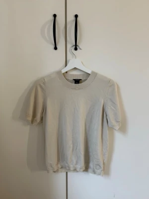 Beige kortärmad topp från Lindex - En enkel och stilren beige topp från Lindex i storlek M. Toppen har korta ärmar, rund halsringning med ribbad kant och är gjord i ett mjukt material som känns skönt mot huden. Perfekt att matcha med jeans eller kjol för en clean look.