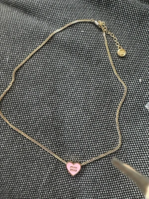Guldhalsband med rosa hjärthänge - Snyggt halsband i guldfärgad metall med ett hjärthänge i rosa emalj där det står 'miu miu'. Kedjan är tunn och har ett litet runt märke vid låset. Baksidan av hänget är guldigt och märkt med 'MADE IN ITALY'. Perfekt accessoar för att lyfta din outfit.