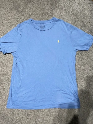 Blå t-shirt från Polo Ralph Lauren - Snygg blå t-shirt från Polo Ralph Lauren med klassisk gul logga broderad på bröstet. T-shirten har rund hals och korta ärmar, perfekt för en avslappnad stil. Tillverkad i mjuk bomull för skön komfort.