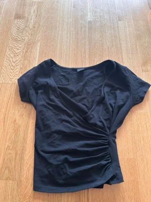 Svart omlottopp med kort ärm - Svart omlottopp i mjukt bomullstyg med korta ärmar och snygg rynkning i sidan. Toppen har en djup v-ringning och sitter figurnära, perfekt till jeans eller kjol.