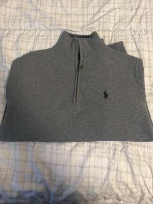 Grå halvzip tröja från Polo Ralph Lauren - Snygg grå stickad tröja från Polo Ralph Lauren med halv dragkedja och hög krage. Tröjan har det klassiska mörkblå logobroderiet på bröstet och ribbade muddar vid ärmslut och nederkant. Perfekt för lager på lager och stilren look. ( Använd 1-2 gånger ) 