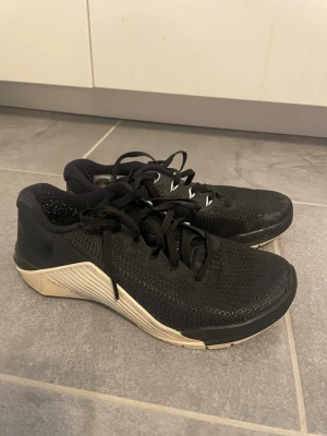 Svarta Nike Metcon 5 sneakers - Säljer ett par svarta Nike Metcon 5 sneakers med vit sula och diskreta detaljer. Skorna har mesh-överdel för bra andningsförmåga och förstärkt häl för extra stabilitet. Perfekta för träning eller en sportig look.