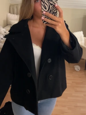 Svart kappa - Säljer en stilren svart kappa med dubbelknäppning och bred krage. Modellen har en klassisk, rak passform. Perfekt att slänga över en hoodie eller tröja för en clean look. Passar till jeans eller kjol för en trendig outfit.