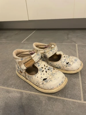 Blommiga vita sandaler för barn - Söta vita sandaler med blått blommönster och kardborreband. Skorna har utskurna detaljer framtill och en mjuk, vadderad insida. Perfekta för vår och sommar, med en klassisk och lekfull design som passar till många outfits.