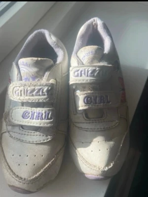 Vita sneakers med kardborre Grizzly Girl(barn)  - Säljer ett par vita sneakers med lila detaljer och texten 'GRIZZLY GIRL' på kardborrebanden. Skorna har lågt skaft och är dekorerade med färgglada mönster på sidorna. Mjukt foder och praktisk kardborrestängning gör dem enkla att ta på. Stolek oklart 