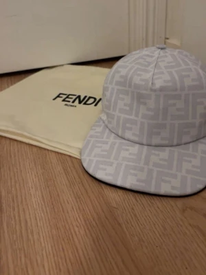 Vit keps med Fendi-logga - Snygg vit keps från Fendi med ikoniskt FF-mönster över hela kepsen. Klassisk form med böjd skärm och justerbar baksida. Tillverkad i tygmaterial och levereras med original dustbag från Fendi.
