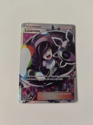 Pokémonkort Lusamine Trainer - Säljer ett Pokémonkort av typen Trainer med motivet Lusamine. Kortet har en färgglad och glansig design med anime-inspirerad illustration och text på engelska. Perfekt för samlare eller Pokémonfans som vill utöka sin kortsamling.